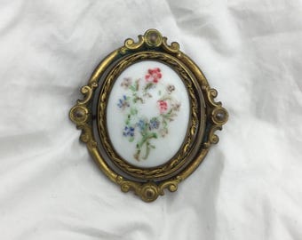 May include: A vintage oval-shaped brooch with a gold-tone metal frame and a white porcelain cameo featuring a hand-painted floral design in shades of pink, blue, and green.