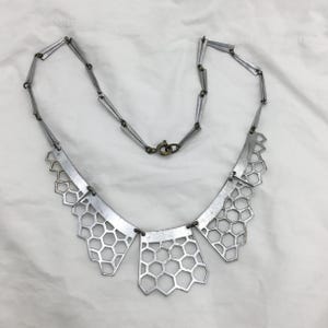 Puede incluir: Collar plateado con un diseño geométrico de panal. El collar presenta una serie de eslabones rectangulares conectados que conducen a un colgante tipo babero. El colgante tiene tres secciones con un patrón de panal y se asegura con un cierre.
