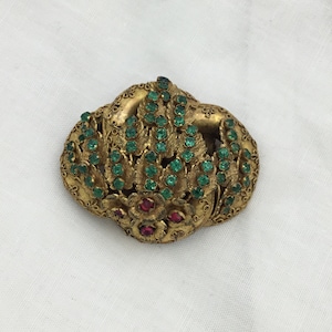 Puede incluir: Un broche de tono dorado con un diseño floral. El broche está adornado con diamantes de imitación verdes y rojos.