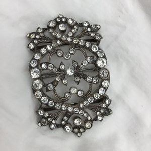 May include: A silver and crystal buckle with an ornate design. The buckle is covered in small, clear crystals that are set in a silver frame. The frame is decorated with intricate swirls and scrolls.