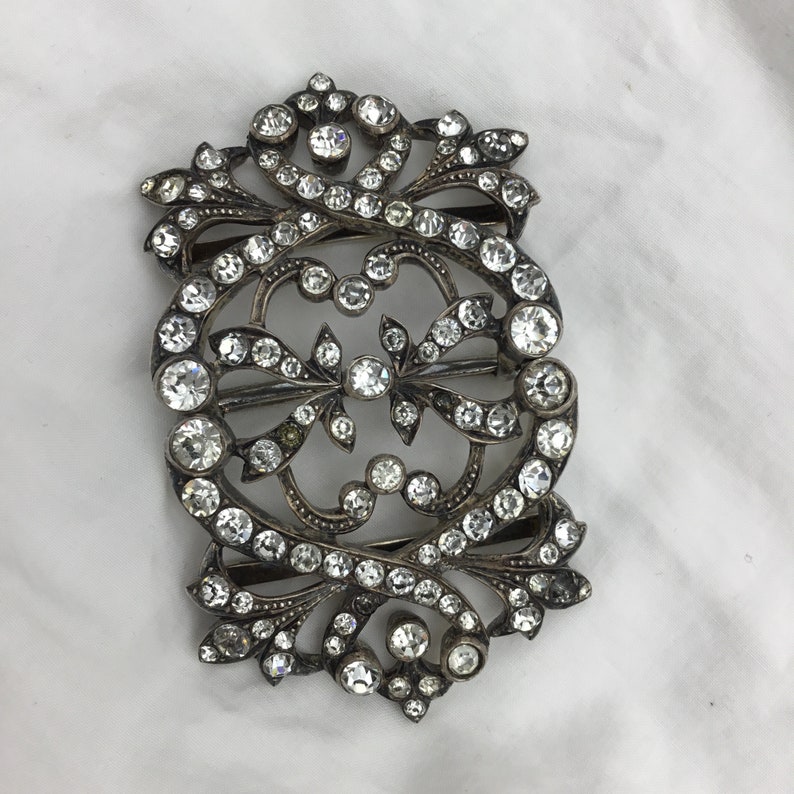 May include: A silver and crystal buckle with an ornate design. The buckle is covered in small, clear crystals that are set in a silver frame. The frame is decorated with intricate swirls and scrolls.