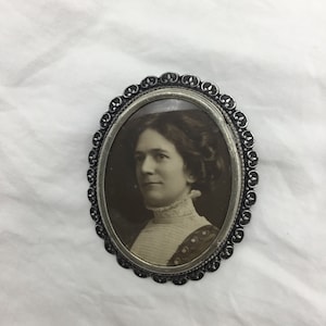 Puede incluir: Un medallón ovalado de plata con una foto en blanco y negro de una mujer en su interior. El medallón tiene un borde decorativo con pequeños diseños de flor de lis.