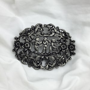 Puede incluir: Broche ovalado plateado con un diseño calado detallado. El centro presenta un relieve de querubines entre elementos florales y foliares. El broche tiene una estética vintage y probablemente está hecho de metal.