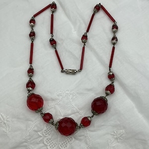May include: A vintage red beaded necklace with faceted and round beads. The necklace features a variety of red beads, including large faceted orbs and smaller round beads, connected by silver-toned metal links and rods. The necklace is displayed on a white surface.