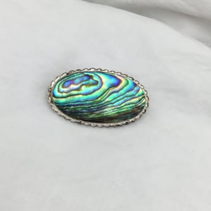 Könnte beinhalten: Eine ovale Brosche aus Silber mit einem irisierenden Perlmutt-Einlegearbeiten aus Abalone. Die Muschel zeigt ein wirbelndes Muster in Blau, Grün und Lila.