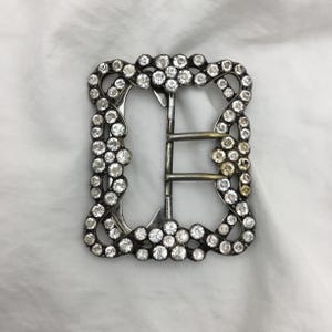 Puede incluir: Una hebilla plateada con un marco decorativo de diamantes de imitación transparentes. La hebilla tiene un cierre de doble lengüeta.