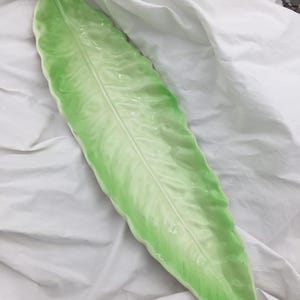 May include: A long, leaf-shaped ceramic serving dish. The dish is a gradient of light and dark green, with a glossy finish. The leaf design has a central vein and detailed texture.
