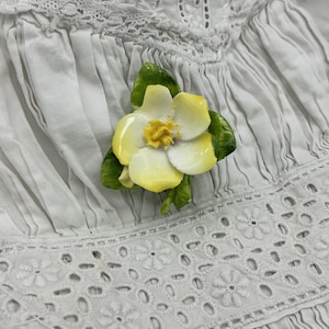 May include: A ceramic flower brooch with white and yellow petals and green leaves. The brooch is set against a white, textured fabric background, suggesting it may be worn on clothing. The flower's center is a bright yellow.