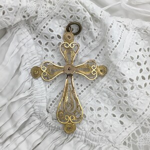 May include: A gold-toned filigree cross pendant with intricate wirework and a circular bail. The cross features a detailed design with spiraled accents and a teardrop-shaped bottom. The pendant is displayed on a white, eyelet fabric background.