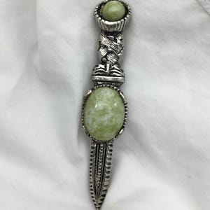 May include: A decorative dagger with a silver-coloured handle and a green, oval-shaped stone in the centre. The handle features intricate detailing and a smaller green stone at the top. The dagger is approximately 20 cm long.