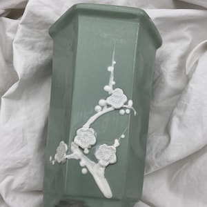 以下が含まれることがあります： セージグリーンの六角形の陶器製花瓶で、白い浮き彫りの梅の花の枝のデザインが施されています。花瓶は滑らかで光沢のある仕上げで、底部の小さな一体型の脚で自立しており、すっきりとした構造的なフォルムを特徴としています。