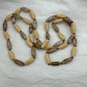 Puede incluir: Un collar largo con cuentas cilíndricas alternas en color crema sólido y patrones de rayas marrones y blancas. Las cuentas están ensartadas en un cordón fino y aseguradas con un cierre de barril cilíndrico de metal tono latón.