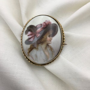 Pu&ograve; includere: Broche ovale in cammeo di porcellana con una montatura dorata. Il cammeo presenta un ritratto dipinto di una donna che indossa un cappello a tesa larga con un nastro rosa.
