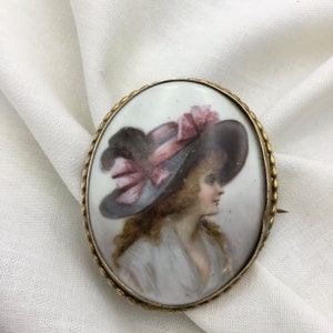 Pu&ograve; includere: Broche ovale in cammeo di porcellana con cornice in metallo dorato. Il cammeo presenta un ritratto dipinto di una donna che indossa un cappello a tesa larga con un nastro rosa.