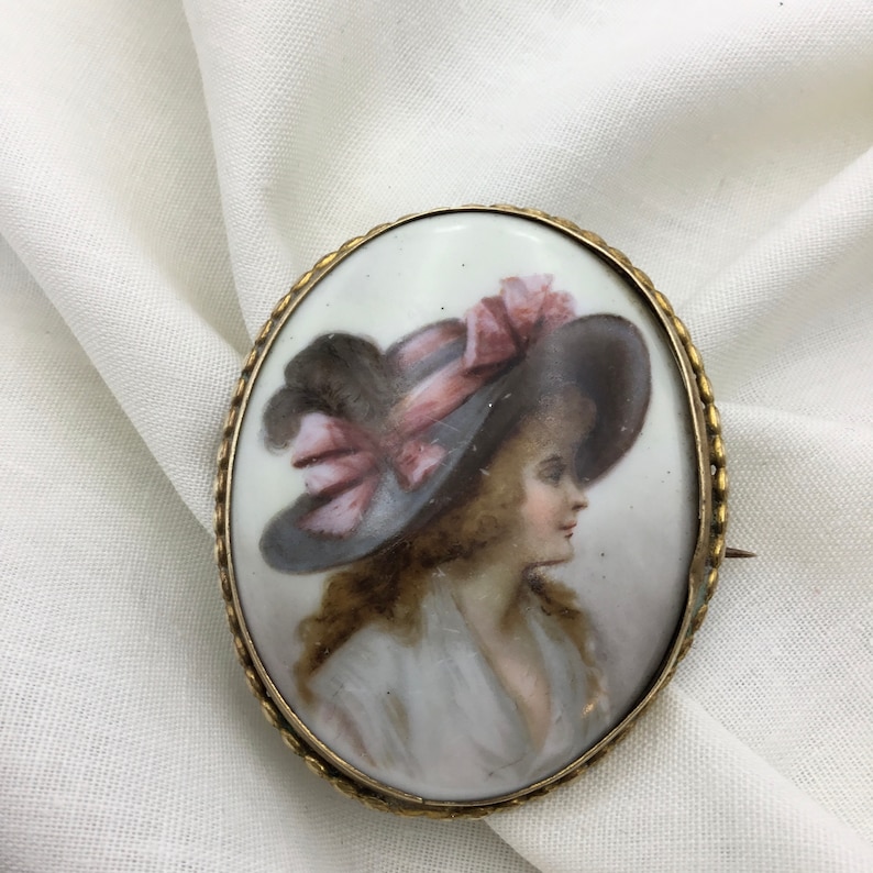 Pu&ograve; includere: Broche ovale in cammeo di porcellana con cornice in metallo dorato. Il cammeo presenta un ritratto dipinto di una donna che indossa un cappello a tesa larga con un nastro rosa.