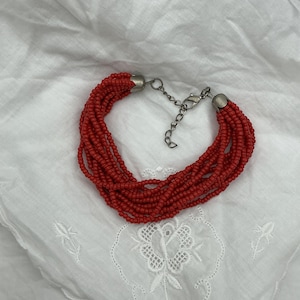 May include: A red beaded bracelet with multiple strands. The bracelet has a silver-colored clasp and end caps. The beads are small and round, and the bracelet is resting on a white, textured surface.