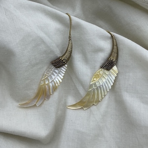 May include: A pair of earrings with a wing design. The earrings feature a gold-colored hook and a decorative section with a spiral wire design. The wings are made of a pearlescent material with a gradient of yellow and white.