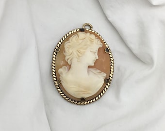 Peut inclure: Un pendentif en camée doré avec un profil sculpté d'une femme dans un camée en coquille. Le camée est serti dans une bordure en forme de corde avec de petits accents dorés.
