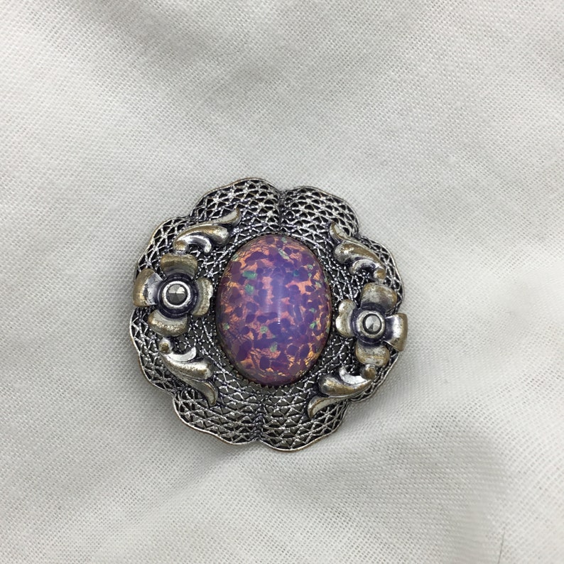 Puede incluir: Un broche de plata con una piedra ovalada iridiscente de color p&uacute;rpura engastada en un dise&ntilde;o de filigrana. El broche tiene dos flores de plata m&aacute;s peque&ntilde;as con centros negros a cada lado de la piedra.