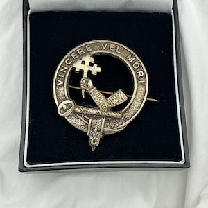 Może przedstawiać: Srebrna okrągła broszka z napisem "VINCERE VEL MORI" i centralnym emblematem dłoni trzymającej krzyż. Broszka umieszczona jest w czarnym pudełku wyłożonym aksamitem. Klasyczny dodatek, prawdopodobnie szkocka odznaka klanu.