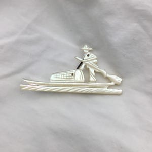 Puede incluir: Broche nacarado que representa una figura en un barco, con sombrero y remo. El barco tiene un compartimento rectangular. El broche está hecho de un material claro, posiblemente nácar, sobre un fondo blanco.