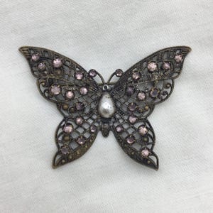 Puede incluir: Un broche de mariposa vintage de bronce con un centro nacarado y un delicado diseño de filigrana. La mariposa está adornada con pequeños diamantes de imitación de color púrpura claro.