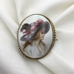 Pu&ograve; includere: Broche ovale in porcellana con cornice dorata. La broche presenta un ritratto dipinto di una donna con un grande cappello con un nastro rosa.
