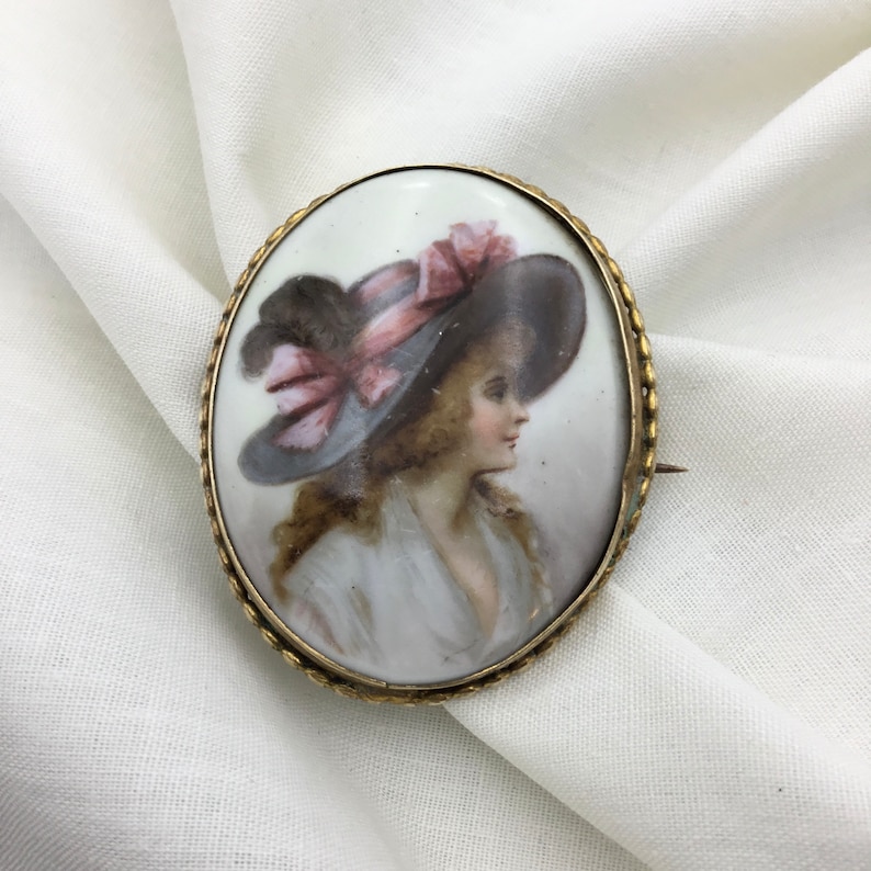 Pu&ograve; includere: Broche ovale in porcellana con cornice dorata. La broche presenta un ritratto dipinto di una donna con un grande cappello con un nastro rosa.