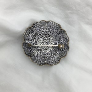 Puede incluir: Un broche de filigrana de plata con un dise&ntilde;o circular y un cierre de pin.