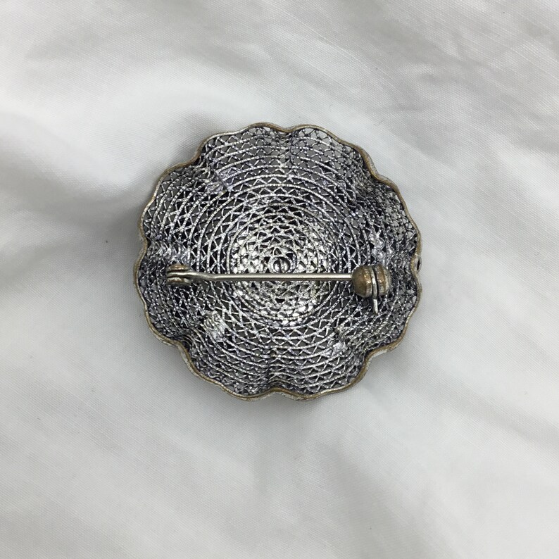 Puede incluir: Un broche de filigrana de plata con un dise&ntilde;o circular y un cierre de pin.