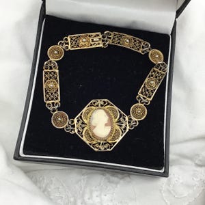 Może przedstawiać: Złota bransoletka w stylu vintage z filigranu z centralnym elementem w postaci kamei. Bransoletka posiada owalny portret kamei w dekoracyjnej ramie, połączony ozdobnymi ogniwami. Bransoletka jest prezentowana w czarnym pudełku.