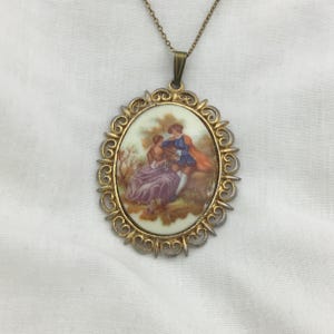 May include: Vintage oval pendant necklace with a painted scene of two figures in a garden. The pendant has a gold-toned filigree frame and a matching chain. The scene features figures in purple, blue, and orange clothing.