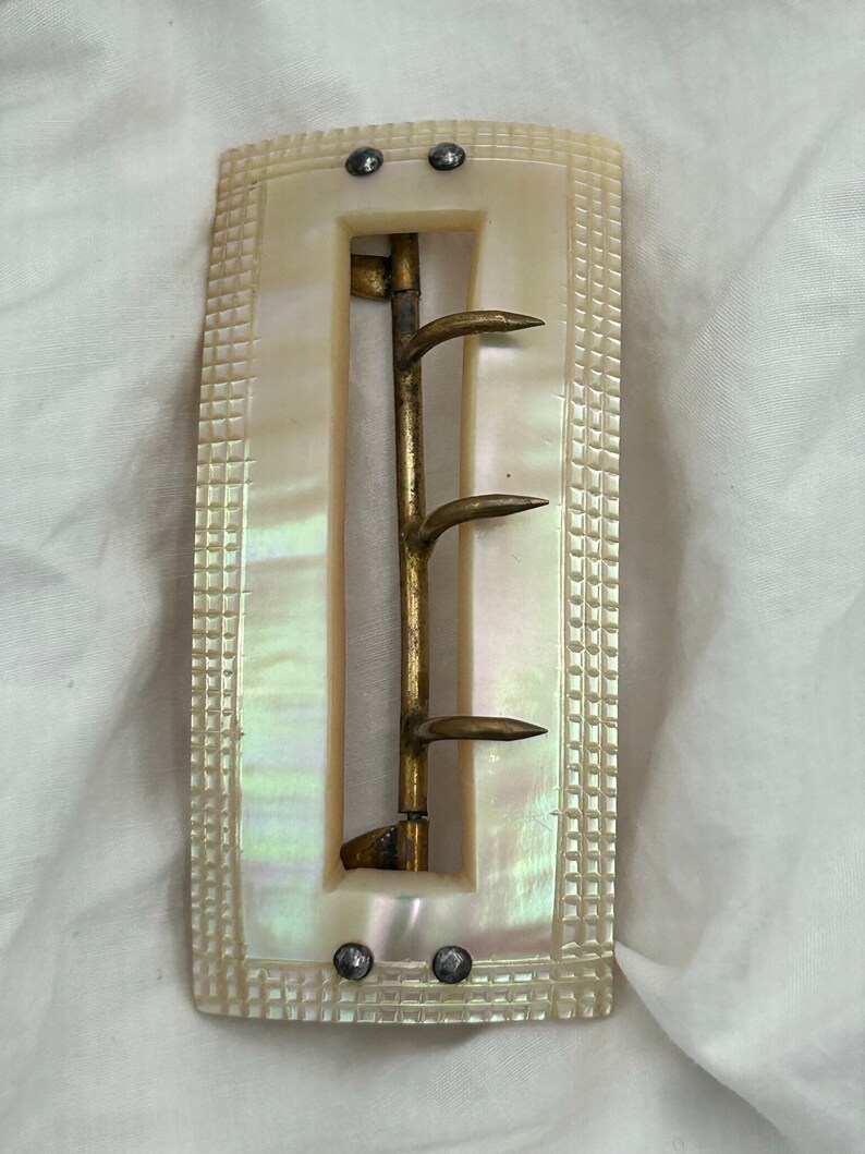 Puede incluir: Una hebilla de cintur&oacute;n de n&aacute;car blanco con un gancho de lat&oacute;n y cuatro puntas. La hebilla tiene una superficie texturizada y es rectangular.