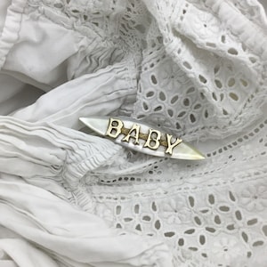 May include: A gold-toned metal pin with the word "BABY" in capital letters, set on a white, elongated, oval-shaped base. The pin is displayed on a white, textured fabric with eyelet details.
