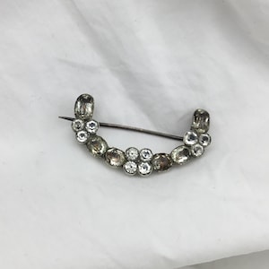 May include: A silver brooch in a curved design, embellished with clear and smoky gemstones. The pin features oval and round stones, creating an elegant accessory. The brooch is set against a white background. A classic piece of jewellery.