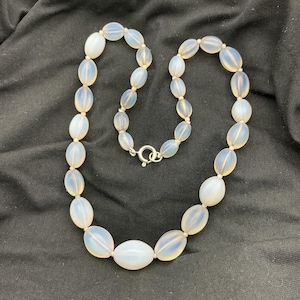 May include: A necklace with oval, opalescent beads strung on a light-coloured cord. The beads are connected with a silver-coloured clasp. The necklace is displayed on a black surface.