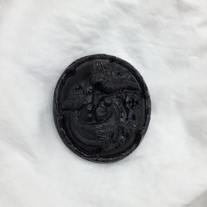 May include: A black glass button with a raised design of two birds perched on a branch with berries. The button has a detailed, intricate design.