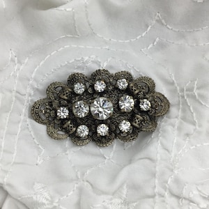 Puede incluir: Broche ovalada de estilo antiguo con diseño de filigrana. La broche presenta brillantes pedrerías transparentes de varios tamaños. El metal tiene un aspecto envejecido y deslustrado. La broche se encuentra sobre una tela blanca estampada.