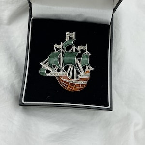 May include: A vintage ship brooch, presented in a black box. The brooch features a detailed sailing ship design with green sails, a brown hull, and silver-tone accents. The brooch is likely made of metal and adorned with small, sparkling embellishments.