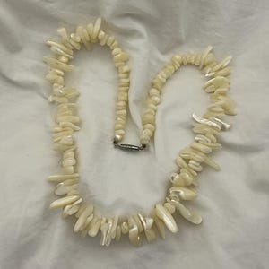 May include: A cream-colored necklace composed of irregularly shaped, polished shell pieces. The necklace features a silver-toned clasp and is displayed on a white, textured surface. The shell pieces vary in size and shape, creating a unique, organic aesthetic.