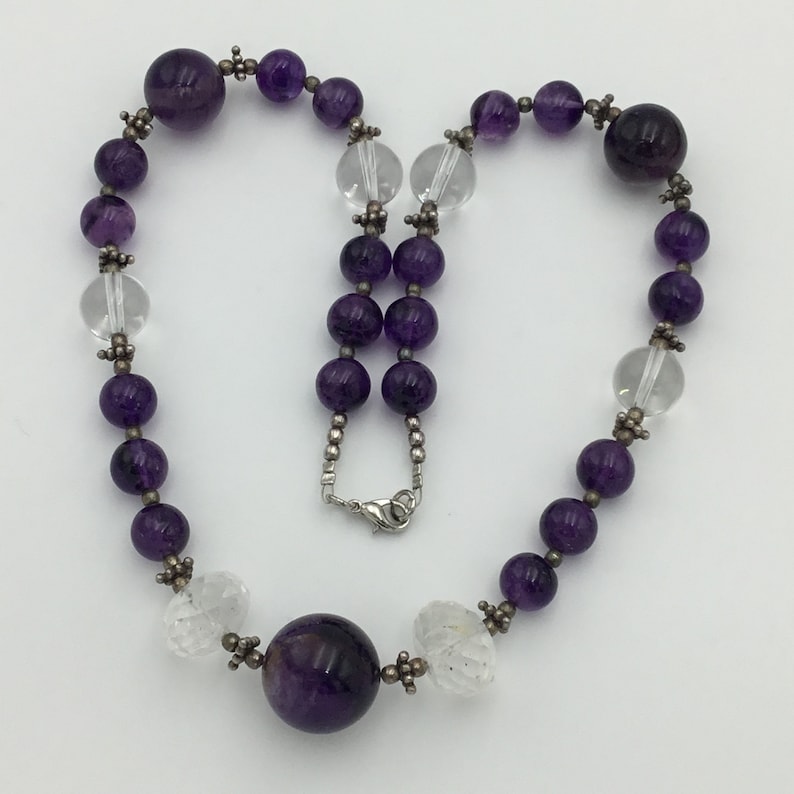 May include: A purple and clear crystal beaded necklace with a silver clasp. The necklace features alternating amethyst and clear quartz beads, with a larger amethyst bead at the bottom.