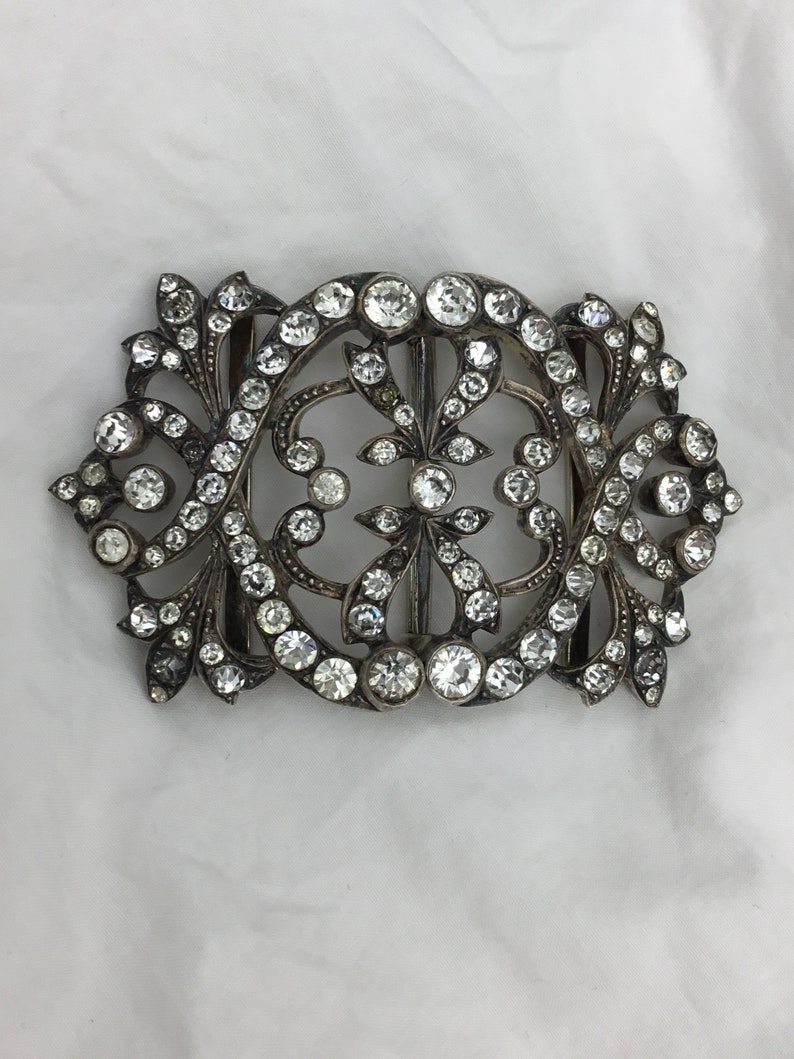 May include: A silver-toned ornate buckle with a floral design, set with clear rhinestones.