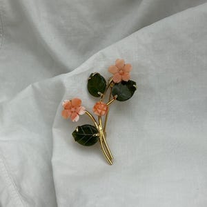 Puede incluir: Broche floral dorado con flores color coral y hojas verdes. El broche tiene un diseño delicado, con las flores y hojas dispuestas sobre finos tallos. Un accesorio clásico.
