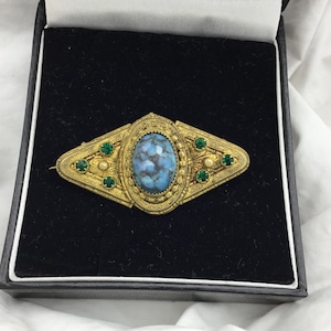 May include: A vintage gold-toned brooch with an oval, speckled blue center stone. The brooch has a diamond-shaped design with green accent stones and intricate detailing, presented in a black jewelry box.