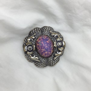 Puede incluir: Un broche de plata con una piedra ovalada de color p&uacute;rpura iridiscente engastada en un dise&ntilde;o de filigrana. El broche tiene dos peque&ntilde;os detalles de flores a cada lado de la piedra.