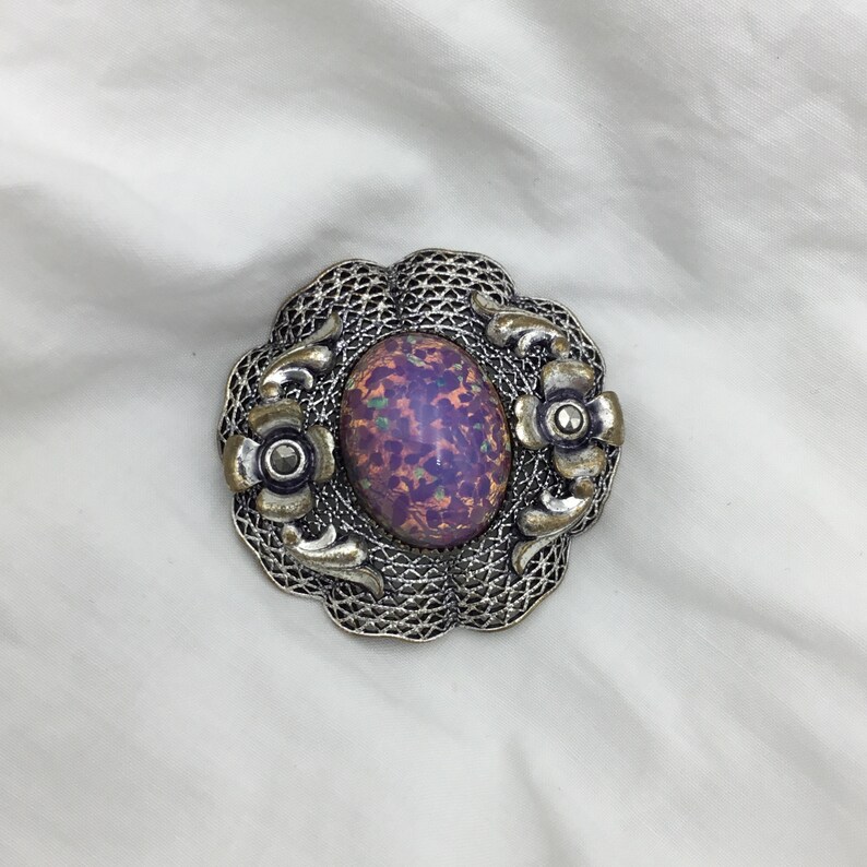 Puede incluir: Un broche de plata con una piedra ovalada de color p&uacute;rpura iridiscente engastada en un dise&ntilde;o de filigrana. El broche tiene dos peque&ntilde;os detalles de flores a cada lado de la piedra.
