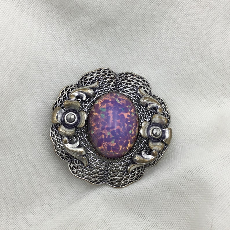 Puede incluir: Un broche de plata con una piedra de &oacute;palo ovalada iridiscente de color p&uacute;rpura y verde engastada en un dise&ntilde;o de filigrana.