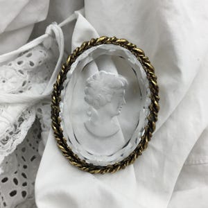 Puede incluir: Un broche de caméo de cristal transparente con un borde dorado en forma de cuerda. El caméo presenta un perfil en relieve de un rostro de mujer.