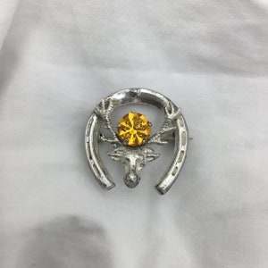 May include: Silver horseshoe brooch with a deer head and a large yellow rhinestone in the center.