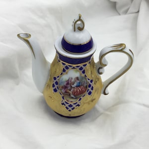 May include: A small porcelain teapot with a blue and gold design. The teapot has a lid with a white knob and a handle on the side. The teapot is decorated with a floral pattern and a scene of two people in a garden.
